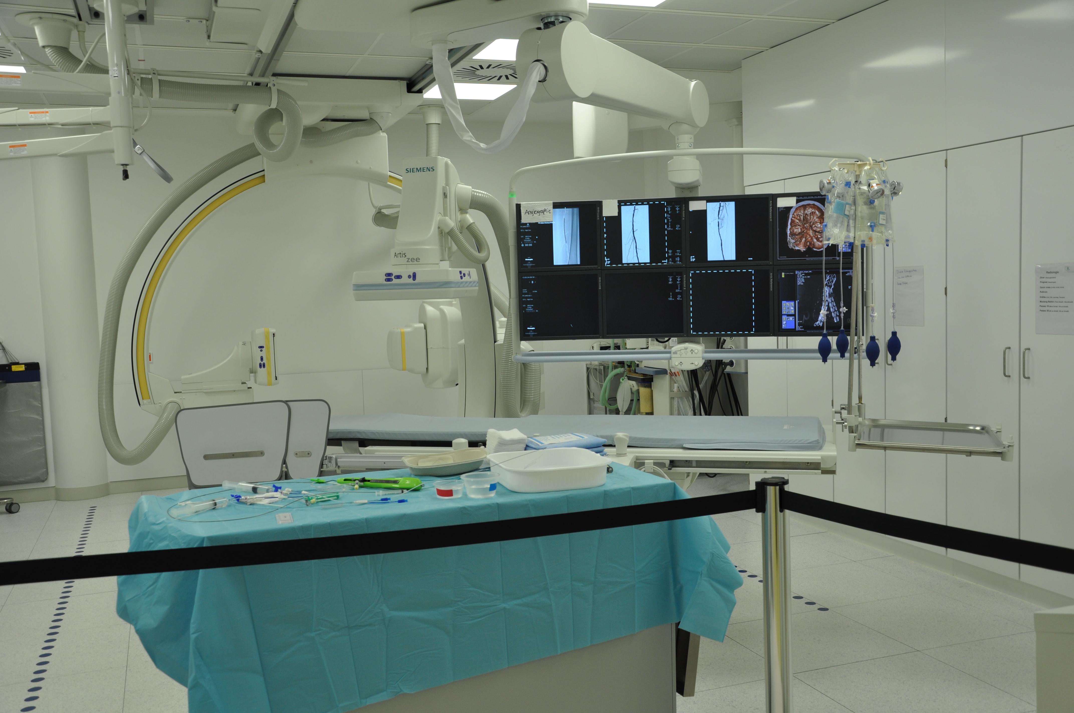 Photograph of a fluoroscopy procedure room with a C-arm X-ray machine, monitor display, and procedure table draped with sterile blue cloths.